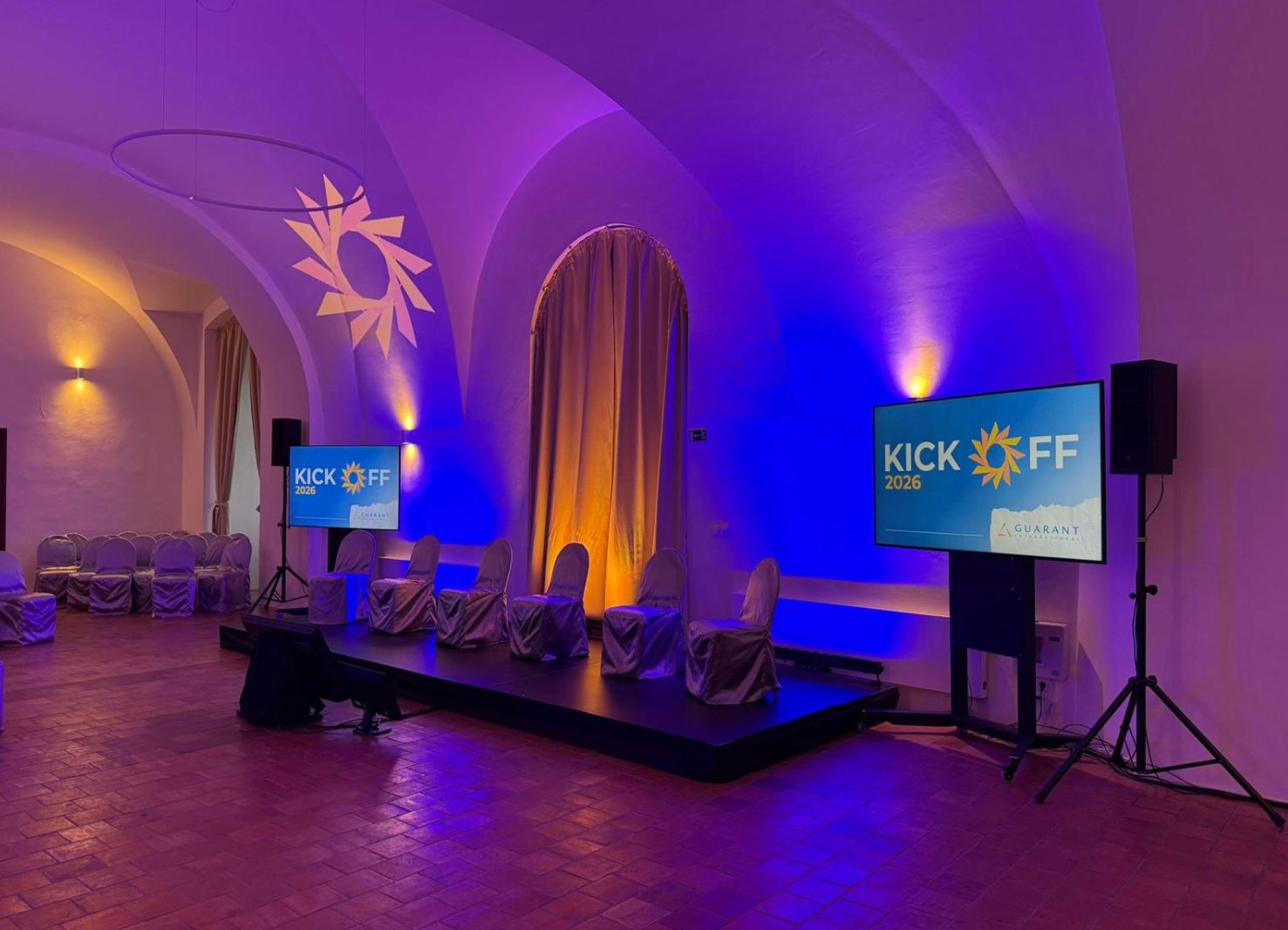 Sál nasvícený do modrofialova, dvě LED obrazovky s logem KICK OFF 2026 a pódium