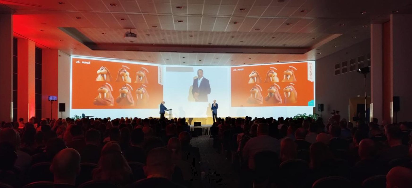 Zaplněný konferenční sál s velkoplošnou projekcí a řečníkem na pódiu